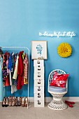 Clothes hanging on rail next to stack of white storage boxes and wicker chair under illuminated neon lettering on wall painted light blue