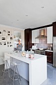 Glossy, white counter with sink and plexiglass stools; fitted kitchen with dark fronts and scattered gallery of photos in background