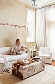 Text artwork on sand-coloured wall above sofa with white loose cover and ottoman with animal-skin cover; woman reading on sofa