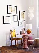 Tisch mit geblümter Tapete beklebt, darauf Vasen und Tischlampe; verschiedene Sitzgelegenheiten vor weisser Wand mit gerahmten Bildern