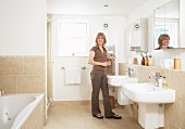 Woman in bathroom with twin sinks below mirrored cabinets and sand-coloured tiles on floor and walls