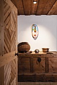 View through open wooden door of modern, painted wall-mounted sculpture and ornaments on carved farmhouse trunk