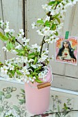 Wild cherry blossom in pink milk can on antiquarian botanical book in front of postcard of the Madonna on white wooden wall
