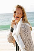 A young blonde woman by the sea wearing a beige knitted cardigan and a scarf