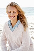 A young blonde woman by the sea wearing a denim shirt and a white woollen jumper