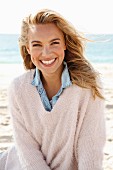 A young blonde woman by the sea wearing a denim shirt and a white woollen jumper