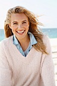 A young blonde woman by the sea wearing a denim shirt and a white woollen jumper