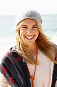A young blonde woman by the sea with a light jumper, a scarf and a hat