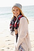 A young blonde woman by the sea wearing a light jumper, a scarf and a hat