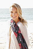 A young blonde woman by the sea wearing a light jumper and a scarf