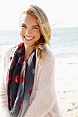 A young blonde woman by the sea wearing a light jumper and a scarf