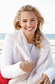 A young blonde woman by the sea wearing a light knitted jumper with another jumper over her shoulders