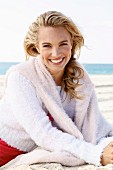 A young blonde woman by the sea wearing a light knitted jumper with another jumper over her shoulders