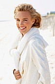 A young blonde woman on a beach wearing a white coat with a jumper over her shoulders