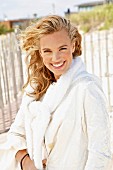 A young blonde woman on a beach wearing a white coat with a jumper over her shoulders