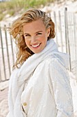 A young blonde woman on a beach wearing a white coat with a jumper over her shoulders