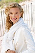 A young blonde woman on a beach wearing a white coat with a jumper over her shoulders