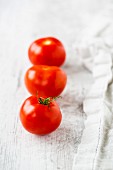 A row of three tomatoes next to a tea towel on a white wooden surface