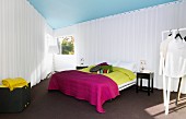 Schlafbereich mit weissen Vorhängen abgetrennt, Doppelbett mit farbigen Tagesdecken auf dunklem Teppichboden in dreieckigem Schlafzimmer, Stehleuchte zwischen Fenstern, im Vordergrund Kleiderständer mit weisser Kleidung