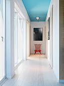 Light-flooded hallway with pale wooden floor, red chair below picture on end wall and spotlight on ceiling painted light blue