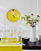 Wanduhr selbstbauen - Ehemaliges Tablett farbig gestrichen mit Uhrwerk ausgestattet, an geweisselter Ziegelwand, davor Glaskaraffe und Weingläser auf gelbem Tischläufer, neben Blumenstrauss in weisser Vase auf schwarzem Tisch