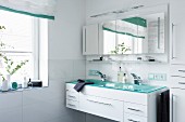 A trough-style washstand for two with a shimmering turquoise glass top built into a white unit with a multi-door, mirrored cabinet hanging above it in a modern bathroom with a window