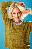 A young blonde woman with her hands behind her head wearing an olive-green knitted jumper