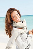 A young brunette woman by the sea wearing a grey jumper and a scarf