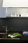 Detail of kitchen counter with stainless steel sink in black worksurface; grey painted glass splashback below open-fronted overhead cabinet with interior light