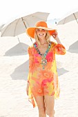 A young blonde woman on a beach wearing a necklace, a summer hat and a long blouse