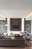 Elegant sofa set in various shades of brown, flatscreen TV on wall in niche flanked by narrow vertical windows and indirect lighting from ceiling
