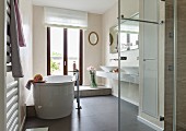 Zeitgenössisches Badezimmer mit freistehender Badewanne vor Balkontür, im Vordergrund verglaster Duschbereich