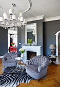 Armchairs with grey velvet upholstery on zebra-skin rug and elegant chandelier in front of mirror above fireplace