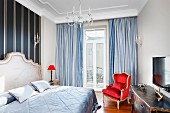 Pale blue blanket on double bed with curved headboard, bedside lamp with red lampshade and balcony door with floor-length curtains