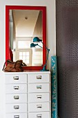 Mirror with red frame and retro table lamp on white chest of drawers