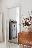 Bulbous vase of flowers on retro sideboard next to open door showing old wooden cupboard in ensuite bathroom