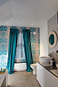 Fenster mit bodenlangem Vorhang in floral gemusterte Tapetenwand, seitlich ein Waschtisch mit Aufsatzbecken auf Unterschränken