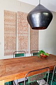 Kunsthandwerkliche Pendelleuchte über Esstisch und Metallstühle mit Patina; Folklore Wandbehänge im Hintergrund