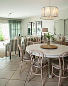 Pendant lamp above dining area with round, white table and Thonet chairs painted pale grey; lounge area in background in bright, modern interior