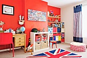 Kinderzimmer mit roter Wand und Teppich mit Union-Jack-Flagge, seitlich Schreibtisch aus Holz, in Nische Regaleinbau mit farbigen Aufbewahrungsboxen