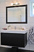 Washstand with black base unit mounted on white tiled wall below framed, illuminated mirror
