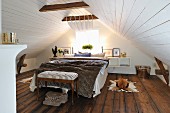 Double bed and antique bedroom bench under white-clad gable ceiling in attic room with dark wooden floor