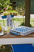 An old board game and a cold drink on a table in a garden with a blue cushion and a maritime t-shirt on garden chairs