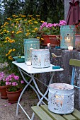 Candles in metal buckets with cut-out designs as lanterns on a table and a chair in a garden