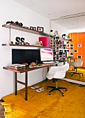 White shell chair at narrow desk below wall-mounted shelves in room with yellow carpet and mirrored wall