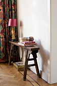 Table lamp with pink lampshade on narrow, antique wooden bench against white wall