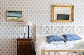 Double bed with white wooden headboard in rustic bedroom with picture and gilt-framed mirror on white and blue patterned wallpaper