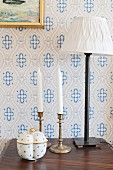 Ceramic pot in front of white candles in candlesticks and table lamp on bedside cabinet against white and blue patterned wallpaper