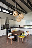 Wooden chairs with colourful backrests and seats around table in white fitted kitchen with spherical pendant lamps