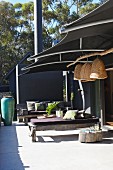 Rustikale Holzbänke mit Liegepolster auf moderner Terrasse, darüber Sonnenschutz aus dunklem Stoff und aufgehängte Stroh Lampenschirme